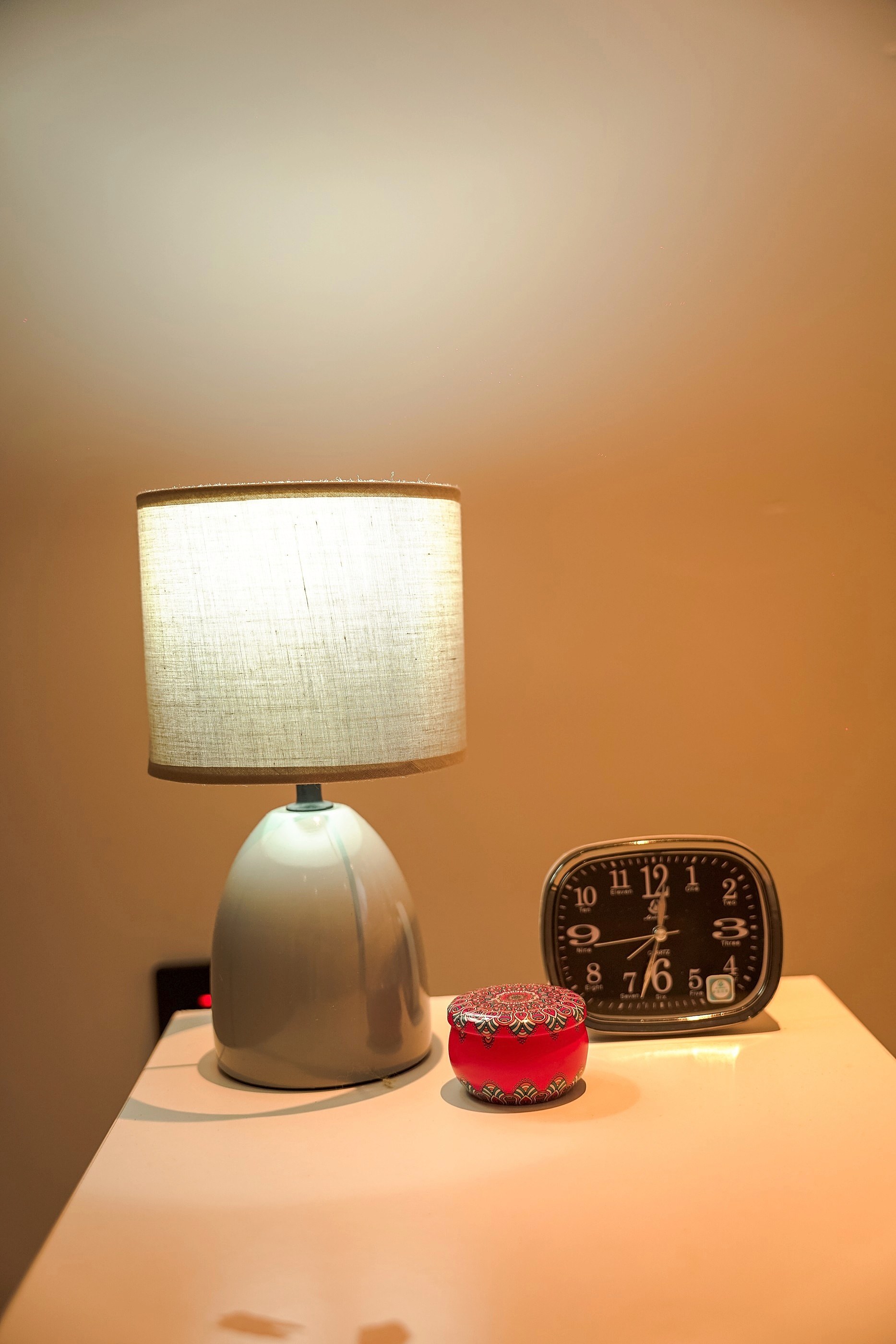 A grey bedside lamp beside an alarm clock and a candle